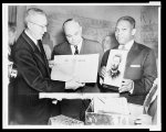 [Hulan Jack holding a photograph of Joseph Pulitzer, while Carl Ackerman and Anthony Donargo look at the cover copy of an 1889 edition of The World (newspaper)]