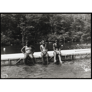 Two girls and a boy sit on a dock with their legs in the water