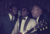 Marion Sledge, Chico Jenkins, and Bobby Moore singing into a microphone on stage at the Laicos Club in Montgomery, Alabama.