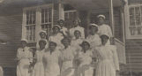 Young African American women holding up baked goods while standing on the steps outside a wooden school building in Jefferson County, Alabama.