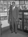 Ralph Metcalfe standing in front of team pictures and athletic trophies, 1932