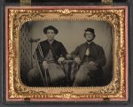 [Two unidentified soldiers in Union private's uniforms sitting next to table with cannon ball on top; one soldier has an amputated leg and holds crutches]