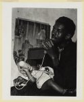 [African American man speaks into a handheld microphone while holding a baby drinking from a baby bottle on his lap at a store front church in Buffalo, New York]