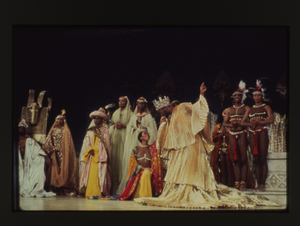 Performance slide of a man in gold wearing a gold crown with a woman wearing red and gold surrounded by various costumed actors]. [color slide