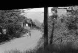 Children walk up a dirt road near a row of houses under a railroad trestle.