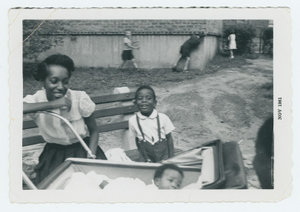 Woman with small boy and baby in a carriage outside on a lawn.