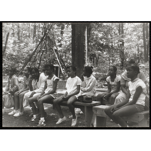 A Group of girls sitting on benches in a wooded clearing at camp