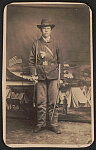 [Private John Cave of Co. C, 2nd Mounted Rifles New York Cavalry Regiment in uniform with sword in front of painted backdrop showing military camp]