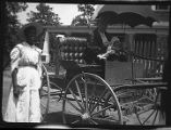 Man sitting in a horse-drawn carriage, holding a baby, with woman standing by the carriage' John Dowe Collection