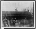 [African American man standing at tall picket fence in front of house]