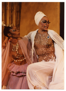 Performance picture of Eartha Kitt (in white turban) with arm on shoulder of woman in rose colored outfit]. [color photoprint