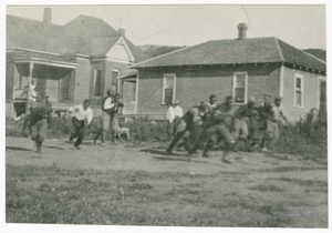 Photograph of men running with a football in front of two houses