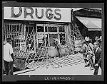 [African American boys and men walking by a drugstore with smashed windows and ripped down gates, on Lenox Avenue, after riots in Harlem, New York City]