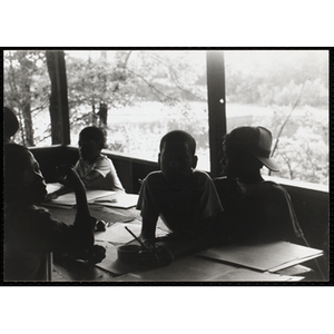 Four boys sit at a table with pencils, paper and scissors