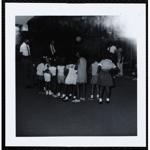 Eight girls, facing away from the camera, stand in a line in front of two male staffers and look at something during a Roxbury Boys' Club event