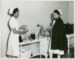 Thumbnail for Captain Mary L. Petty, Chief Nurse, holding a glass bottle and showing it to 2nd Lieutenant Olive Bishop, who is writing on a small pad of paper