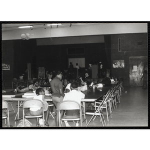 Several Roxbury Boys and Girls Club members sit and stand around tables in an auditorium while others stand on the stage in the distance