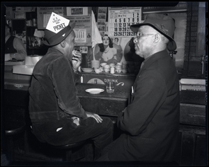 Two men at restaurant counter, one wearing "picket" sign in hat, photonegative