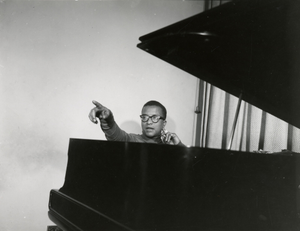 Billy Strayhorn, seated at piano, pointing : black-and-white photoprint.