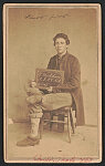 [Private Lamont Wickham of Co. A, 8th New York Heavy Artillery Regiment in uniform with pants rolled up to display his wounded leg, and holding a slate board with his name, regiment, and number 20164]