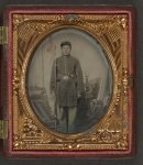 [Unidentified soldier in Union uniform with musket, bayonet in scabbard, revolver, and cap box in front of painted backdrop showing military camp scene]