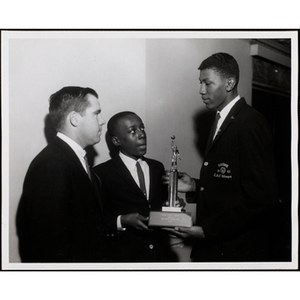 Two young men hold a basketball trophy while another man looks on