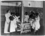 [African American schoolgirls with teacher, learning to cook on a wood stove in classroom]