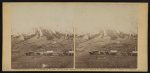 Thumbnail for [Three horse-drawn covered wagons in the foreground.  Soldiers marching in formation between rows of small cabins and tents in the background]