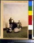 [Four immigrants and their belongings, on a dock, looking out over the water; view from behind]