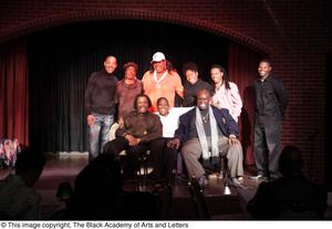 Performer, man, and Curtis King sitting on stage with three women and three men standing