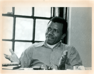 Chinua Achebe gestures while seated at a desk