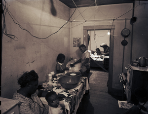 Women and children working and sitting in kitchen, with woman ironing, seen through doorway in background : photonegative.