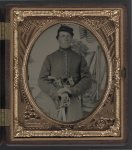 [Unidentified soldier in Union uniform with two Colt revolvers and cavalry sword in front of painted backdrop showing encampment]