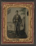 [Unidentified soldier in Union uniform with bayoneted musket, canteen, cartridge and cap boxes in front of painted backdrop showing military camp with American flag]