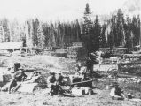 Men cooking and eating while taking a break from their work at the lumber mill seen behind them.