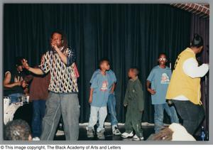 Photograph of a group of children, two men, and a woman performing on a stage