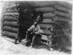 [Afro-American man seated outside log cabin, pouring drink into cup and girl seated in window of the cabin]