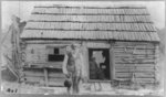 [Stephen Steptoe, Junior, three-quarter length portrait, standing, facing front, in front of cabin with woman in doorway]