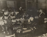 African American man giving a health lecture to an audience of adults and children, probably in a clapboard church building or school.