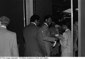 Photograph of three men walking through a doorway while a woman is holding a stack of papers