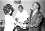 Woman reaching into a paper bag held by a man speaking into a microphone at Tom's Place at 648 South Holt Street in Montgomery, Alabama.