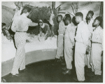 Man pointing to a "Survival Museum" diorama of a soldier sitting under a makeshift bunker in the snow, while African American soldiers from the 25th Regimental Combat Team look on, Pensacola Naval Air Station, Florida