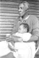 Elderly woman holding a baby while seated on the front porch of a wooden house in Little Korea, a neighborhood in Birmingham, Alabama.