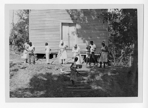 Photograph of African American women and children sweeping and raking land by a wooden building, Clarkesville, Habersham County, Georgia, 1953