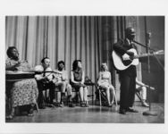 Thumbnail for Mississippi State Sovereignty Commission photograph of an African American male performing on stage with an acoustic guitar and Pete Seeger as well as other musicians participating in the background, Jackson, Mississippi, 1960s