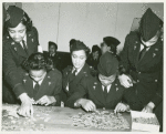 Thumbnail for Two African American Lieutenants of the Women's Army Corps sitting at a table and working on jigsaw puzzles while three others assist and look on