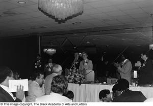 Photograph of a man standing at a podium, while other individuals are standing around and clapping