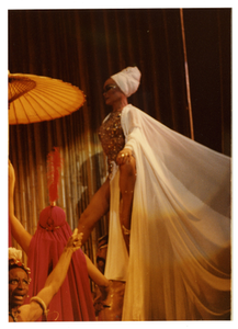 Performance picture of Eartha Kitt in white, one parisol, a woman in the foreground pointing]. [color photoprint