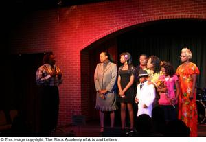 Kirondria Woods, Rachel Webb, and boy in white suit, standing on stage with three women and one man while Curtis King speaks