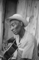 Blues Singers: Lake Mary, Miss. Scott Dunbar playing guitar outside on porch, Martha Dunbar and granddaughter seated outside, interior of Dunbar home (BSP I-68 #130)